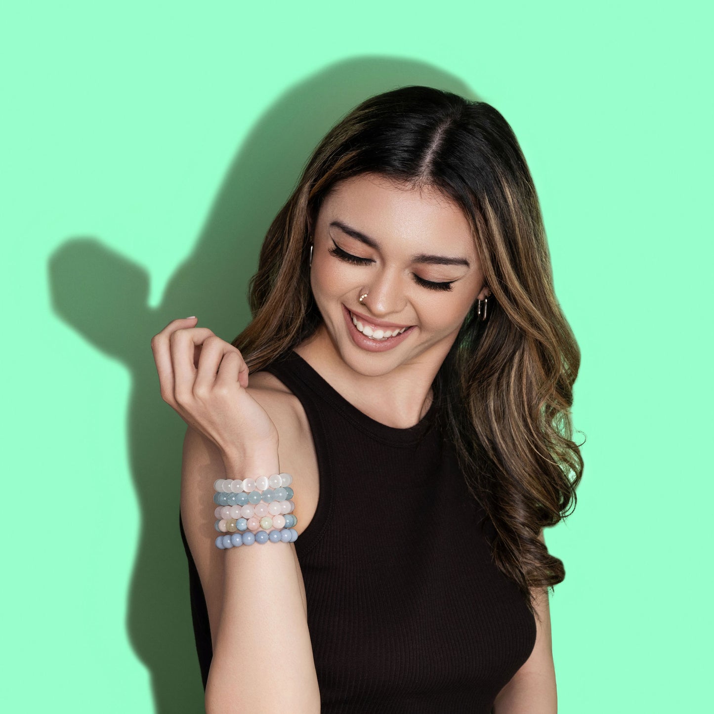 A smiling woman with long hair looks down at a raised wrist. She wears a black top and a large stack of pink, blue, and white crystal gemstone bracelets. The background is bright green.