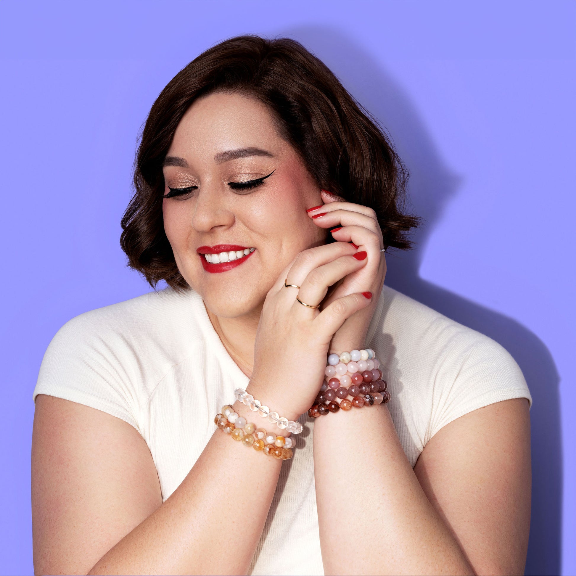 A smiling woman in a white top, with short dark hair and bright red lipstick and nail polish rests her hands to the side of her face. She wears several gold and white gold rings and a stack of crystal gemstone bracelets on each wrist. The background is bright periwinkle.