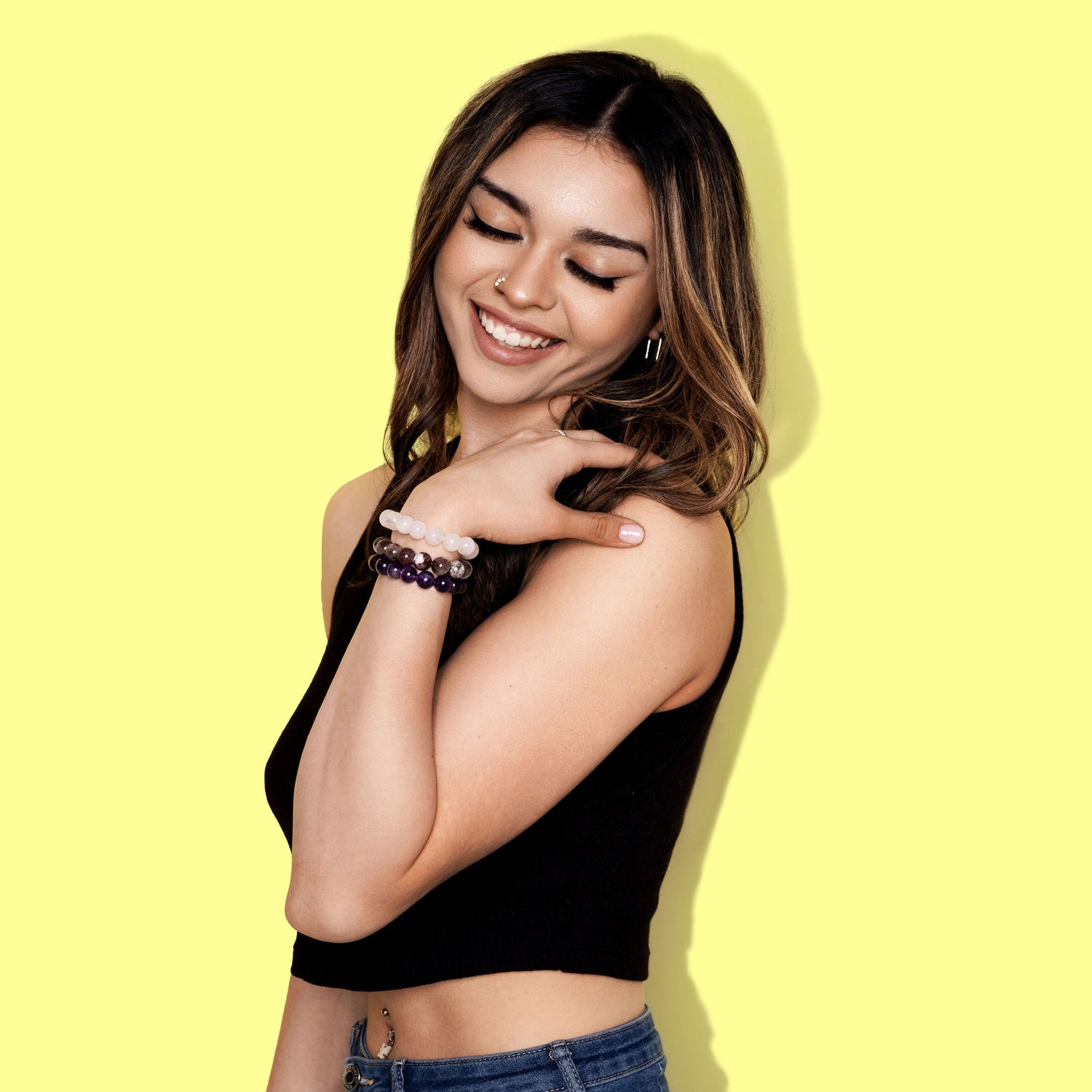 A wide shot of woman with long dark hair wearing a black top and jeans, smiles. She has her gaze cast downwards and rests her hand on her shoulder. She is wearing one pink and two purple coloured crystal gemstone bracelets.