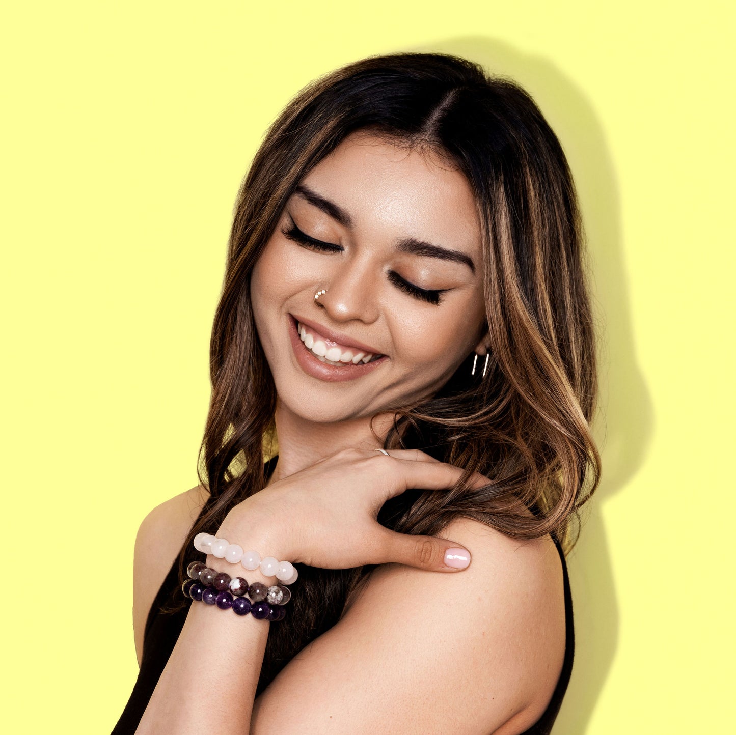 A close up shot of woman with long dark hair wearing a black top, smiles. She has her gaze cast downwards and rests her hand on her shoulder. She is wearing one pink and two purple coloured crystal gemstone bracelets.