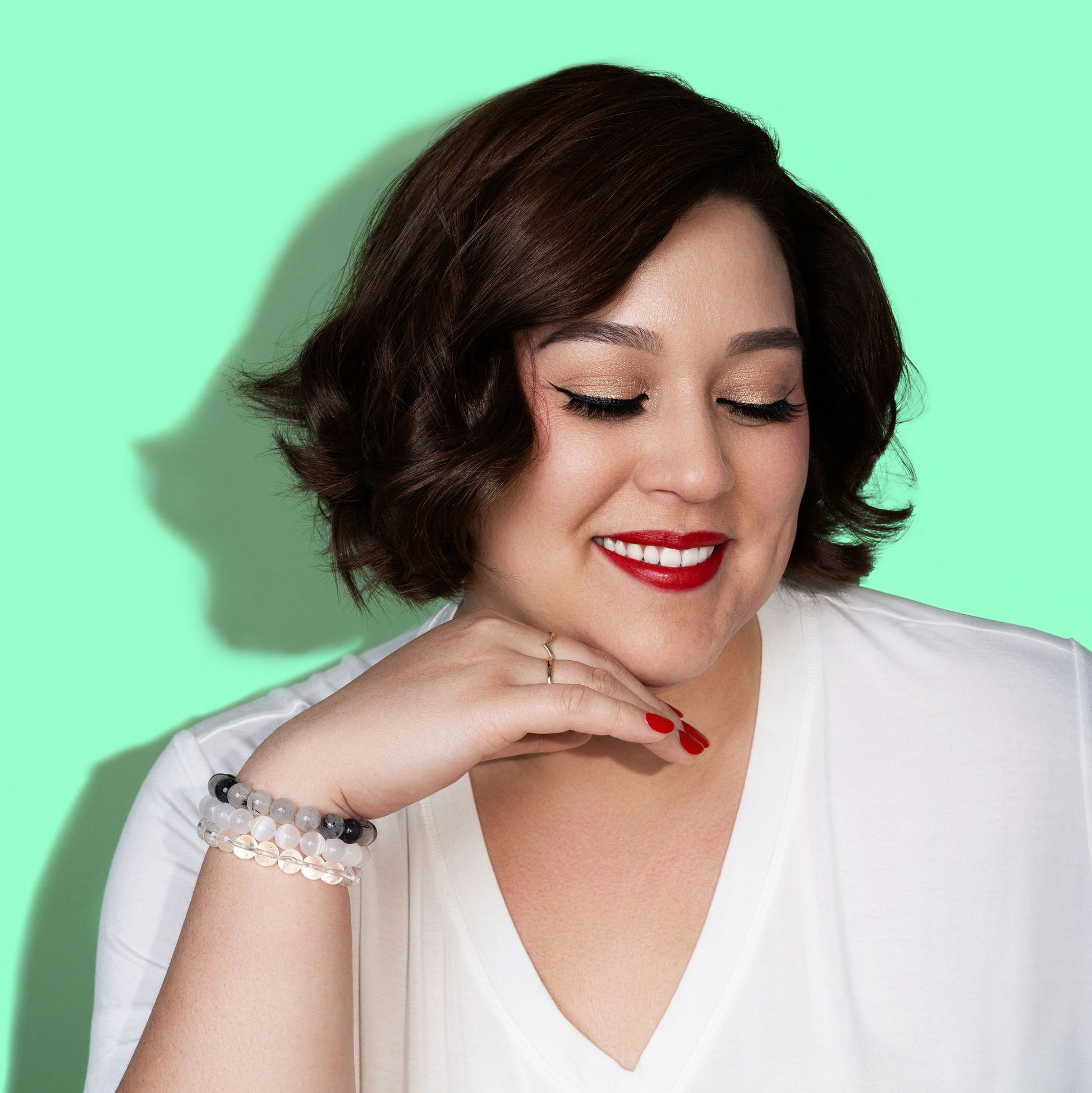 A woman with short dark hair smiles, and wears bright red lipstick. She has her chin on her hand and is wearing three white, clear, and grey coloured crystal gemstone bracelets.