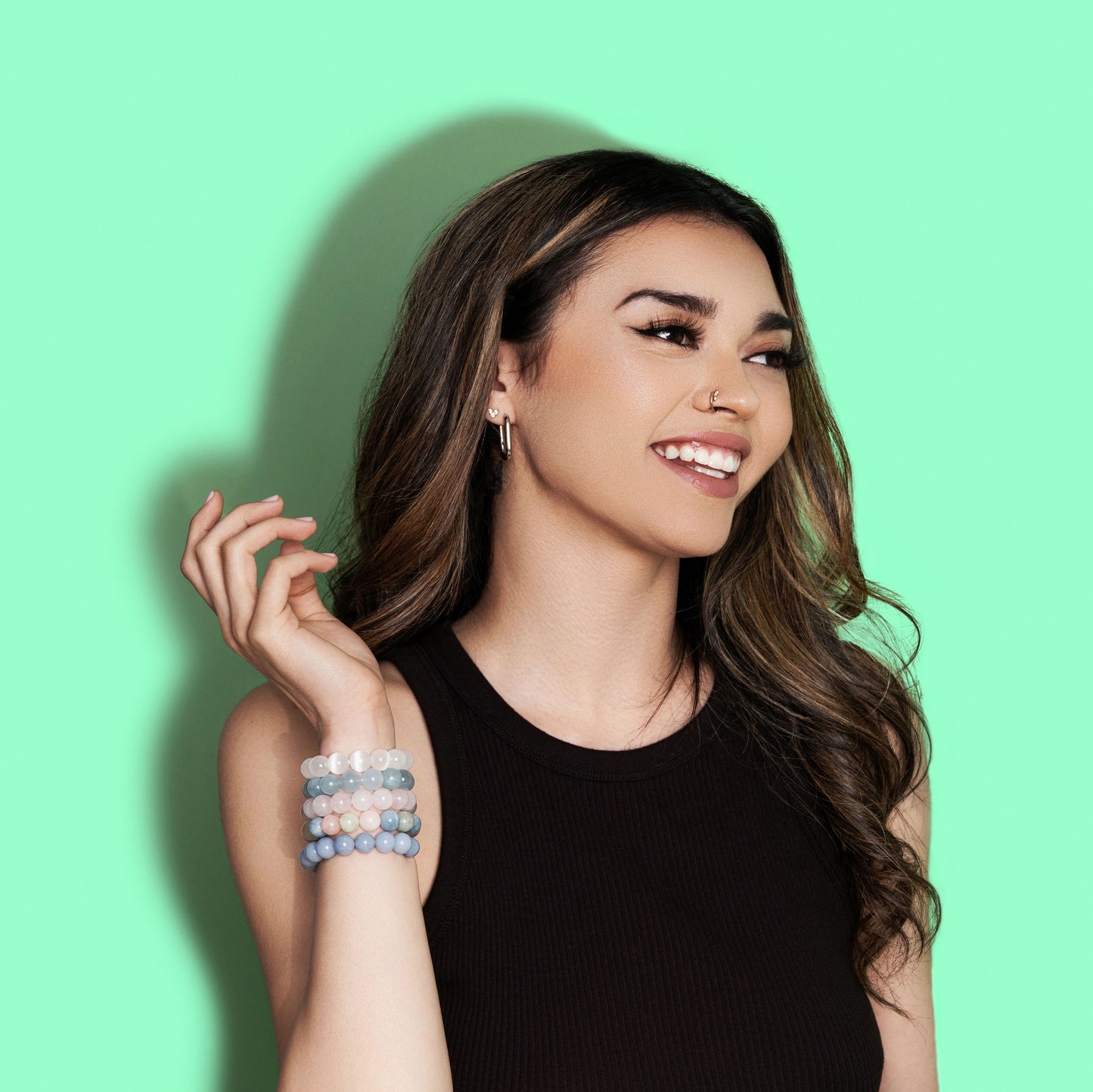 A smiling woman with long dark hair wears a black top and a variety of white, blue, and pink coloured crystal gemstone bracelets. The background is bright green.