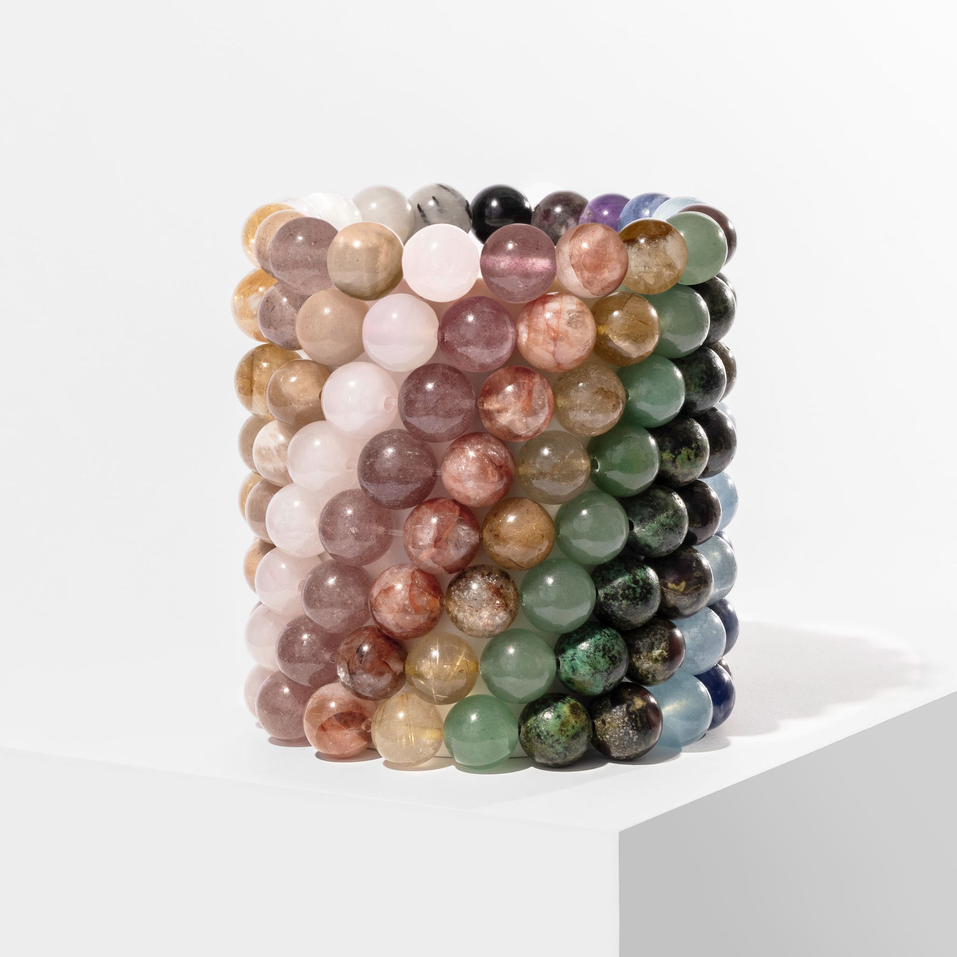 A vertical stack of 8 multicoloured crystal gemstone bracelets, positioned on a white block.