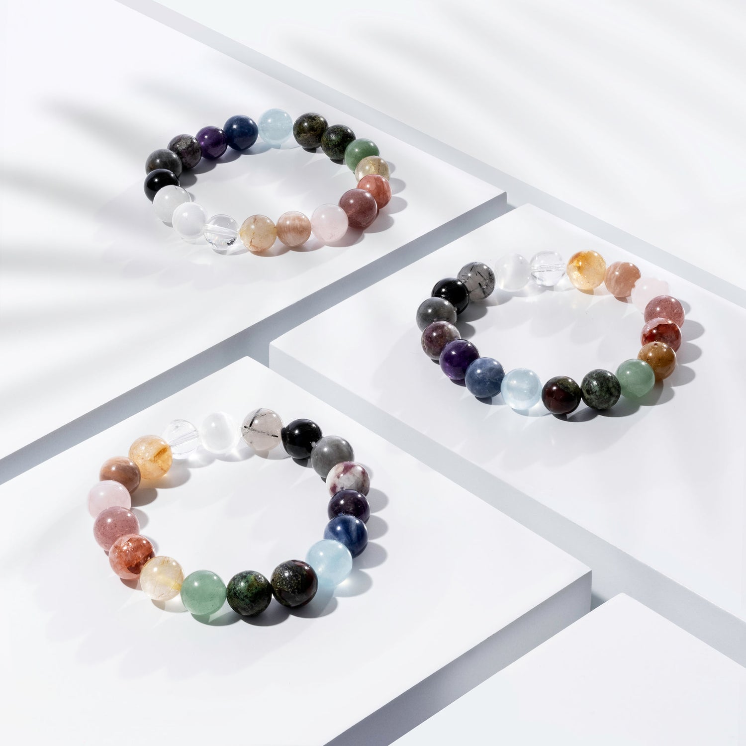 Three multicoloured crystal gemstone bracelets positioned on white blocks. There is a palm foliage shadow dappling light across the display.