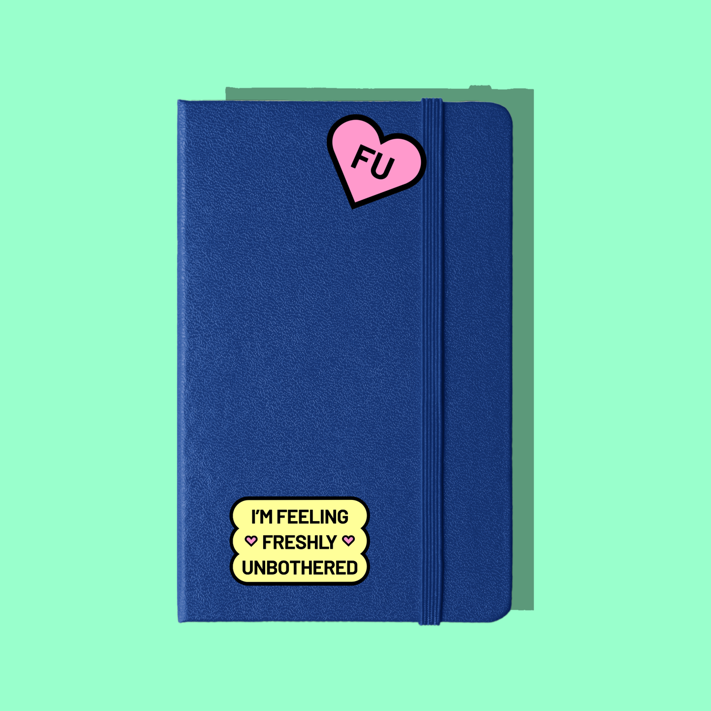 A royal blue notebook has a pink heart and yellow sticker on it. The notebook casts a shadow on a bright green background.