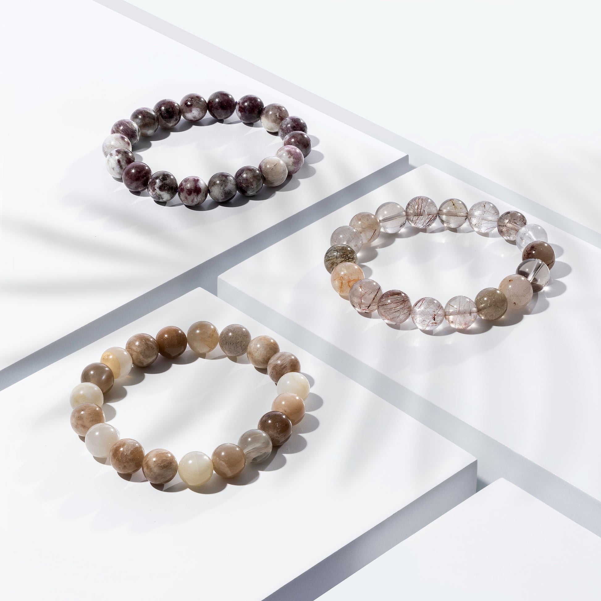 Three crystal gemstone bracelets positioned on white blocks. They are purple, cream, peach, and grey coloured. There is a palm foliage shadow dappling light across the display.