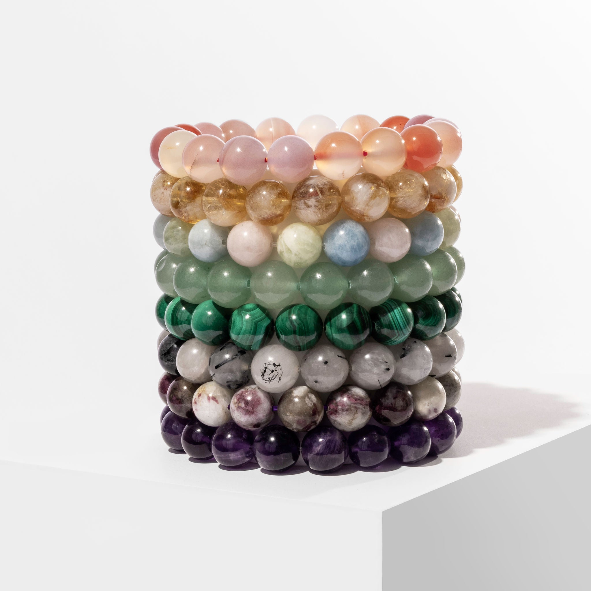 A vertical stack of 8 crystal gemstone bracelets of different colours, positioned on a white block.