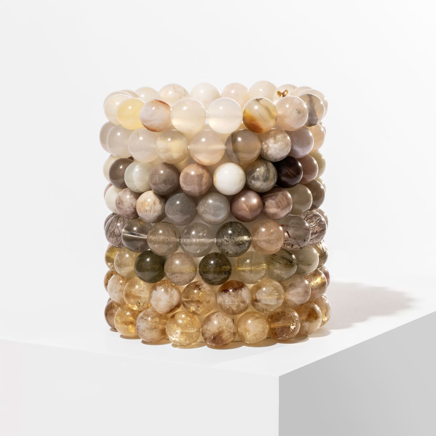 A vertical stack of 8 yellow and cream crystal gemstone bracelets, positioned on a white block.