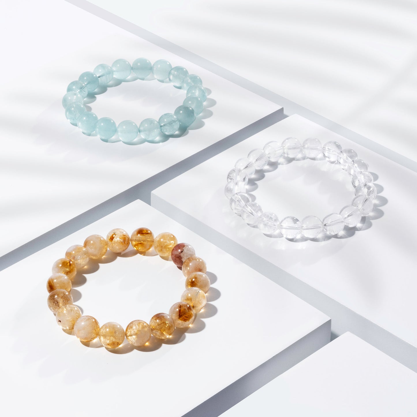 Three yellow, light blue, and clear crystal gemstone bracelets positioned on white blocks. There is a palm foliage shadow dappling light across the display.